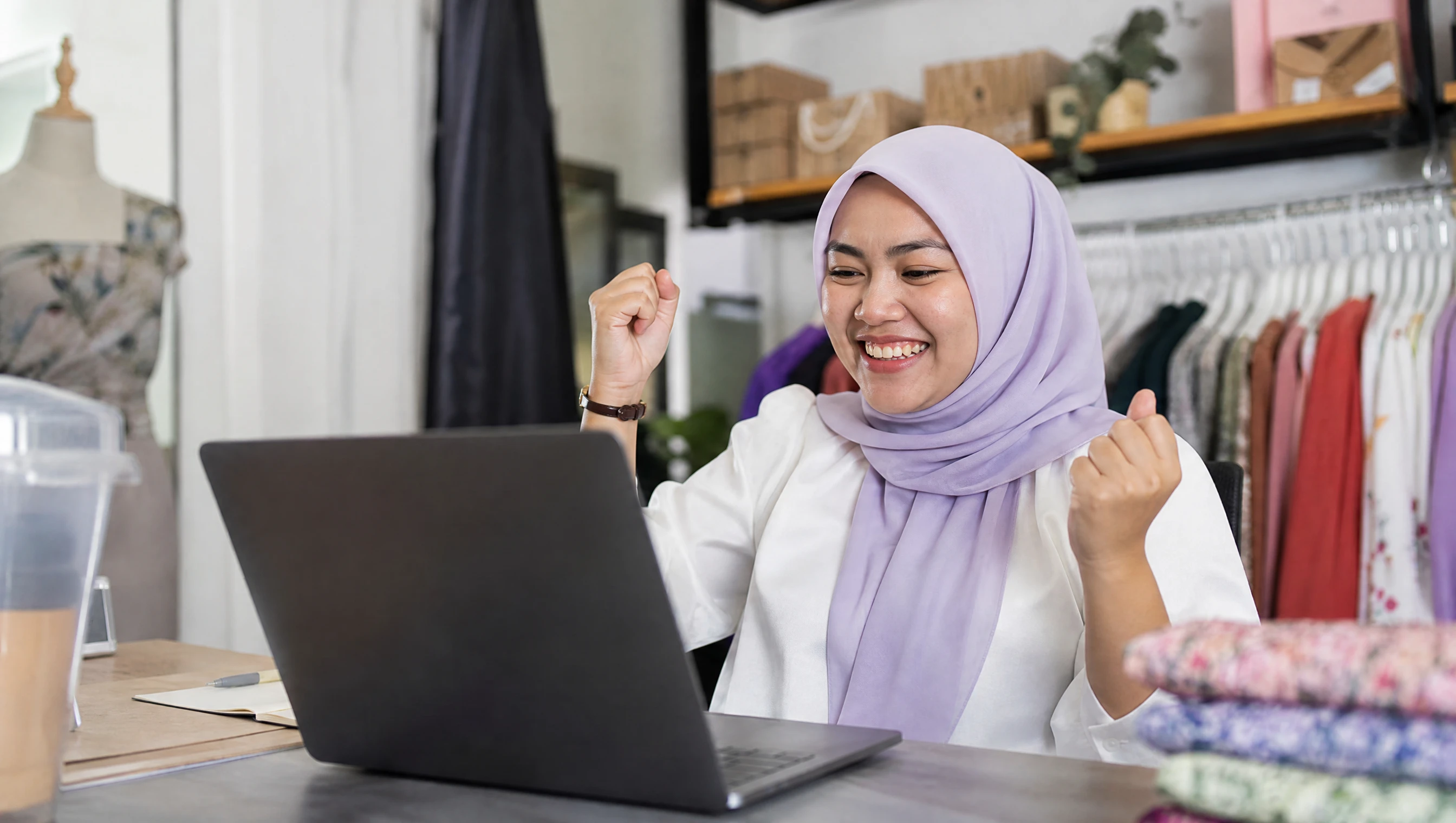 A happy Malaysian business owner in hijab celebrating her newly launched website on a laptop in her shop