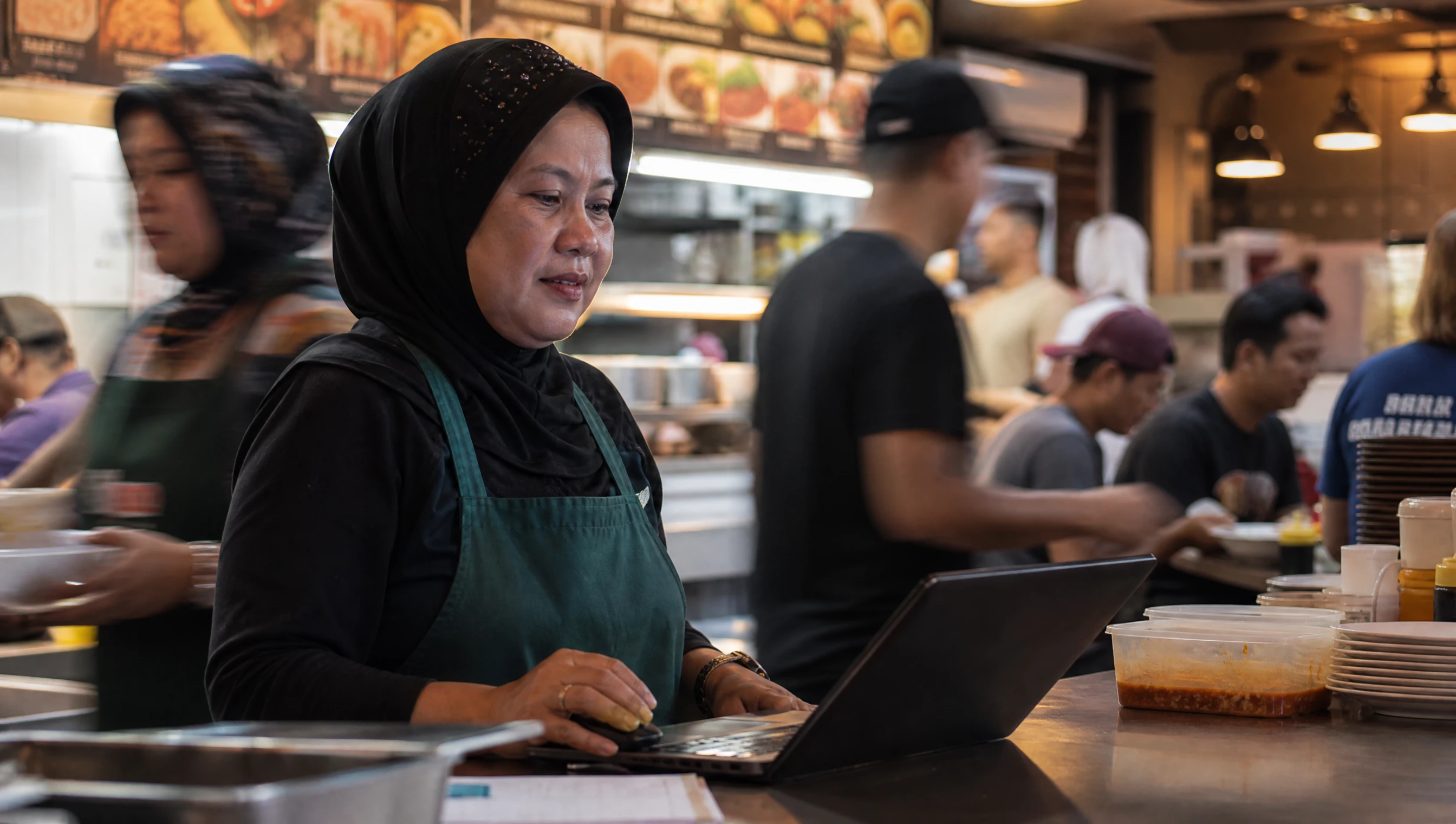 Malaysian restaurant owner in an apron checking a laptop at her counter during a busy lunch service