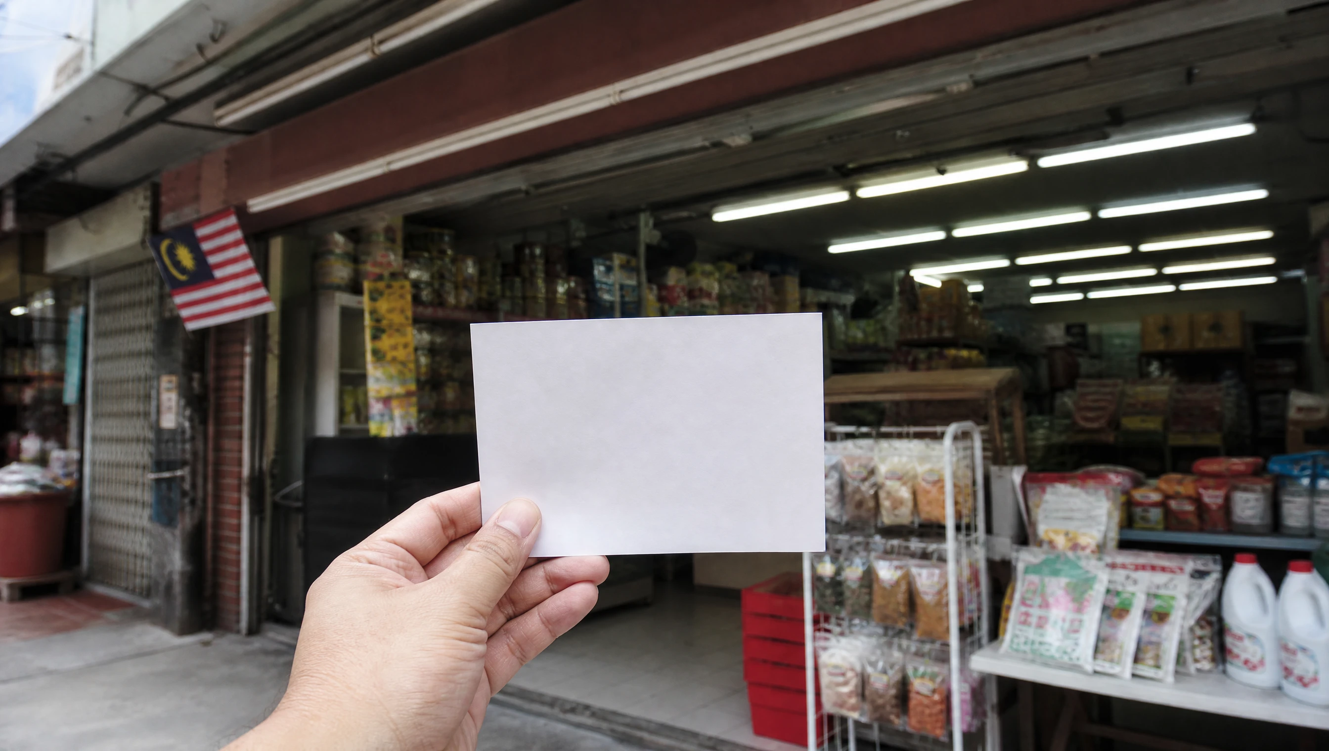 Tangan memegang poskad pengesahan Google di hadapan kedai runcit di Malaysia