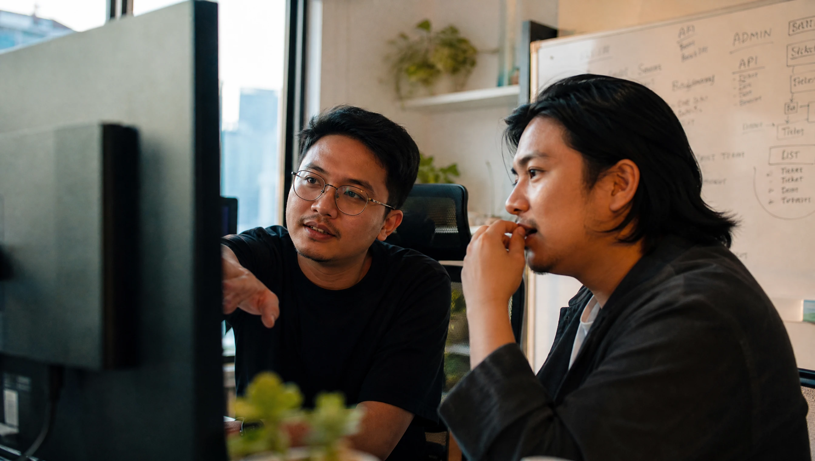 Dua pembangun perisian Malaysia menyemak kod bersama di monitor besar dalam studio kecil di Kuala Lumpur