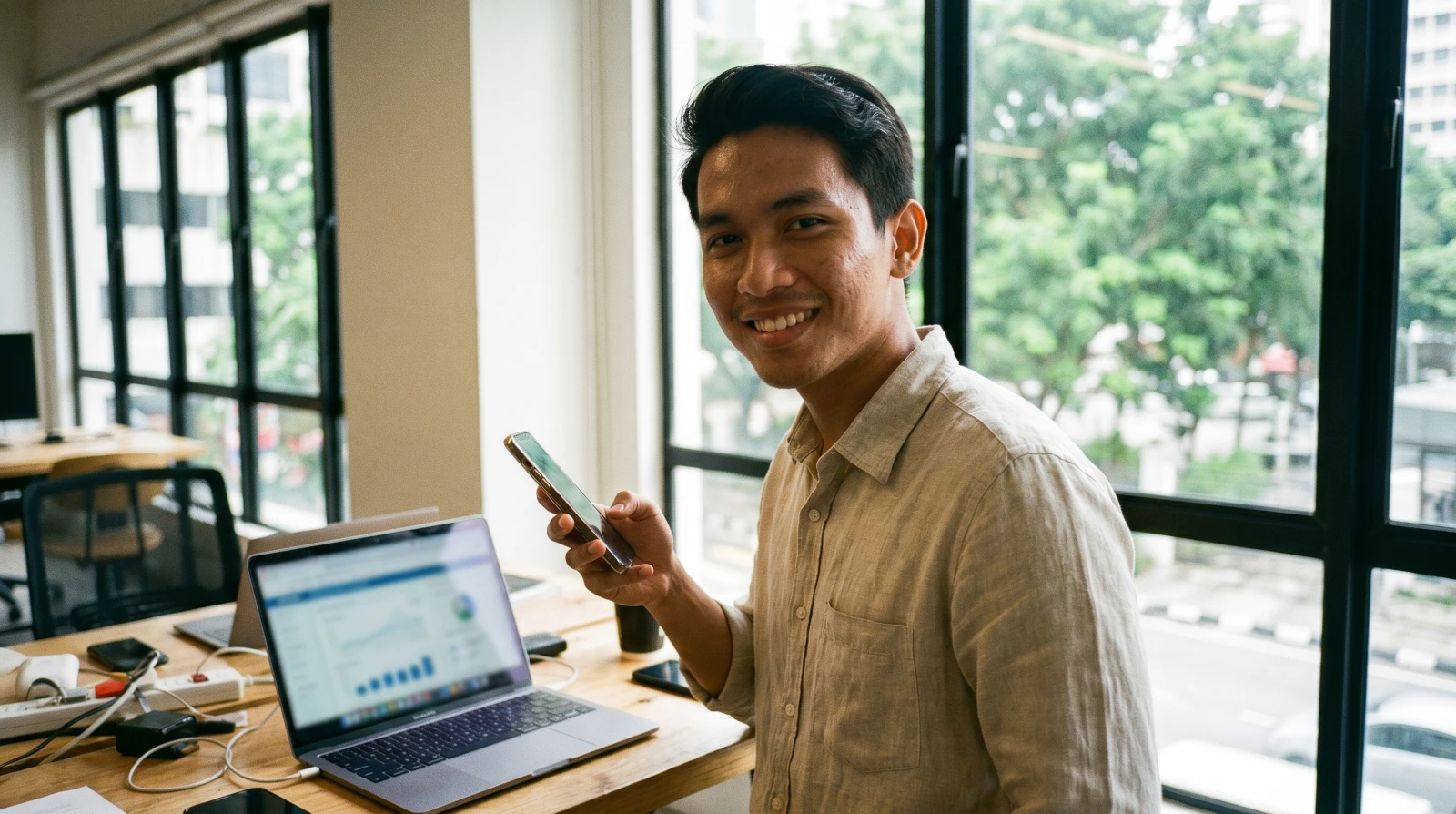 Usahawan lelaki Malaysia tersenyum yakin memegang telefon pintar di ruang kerja moden