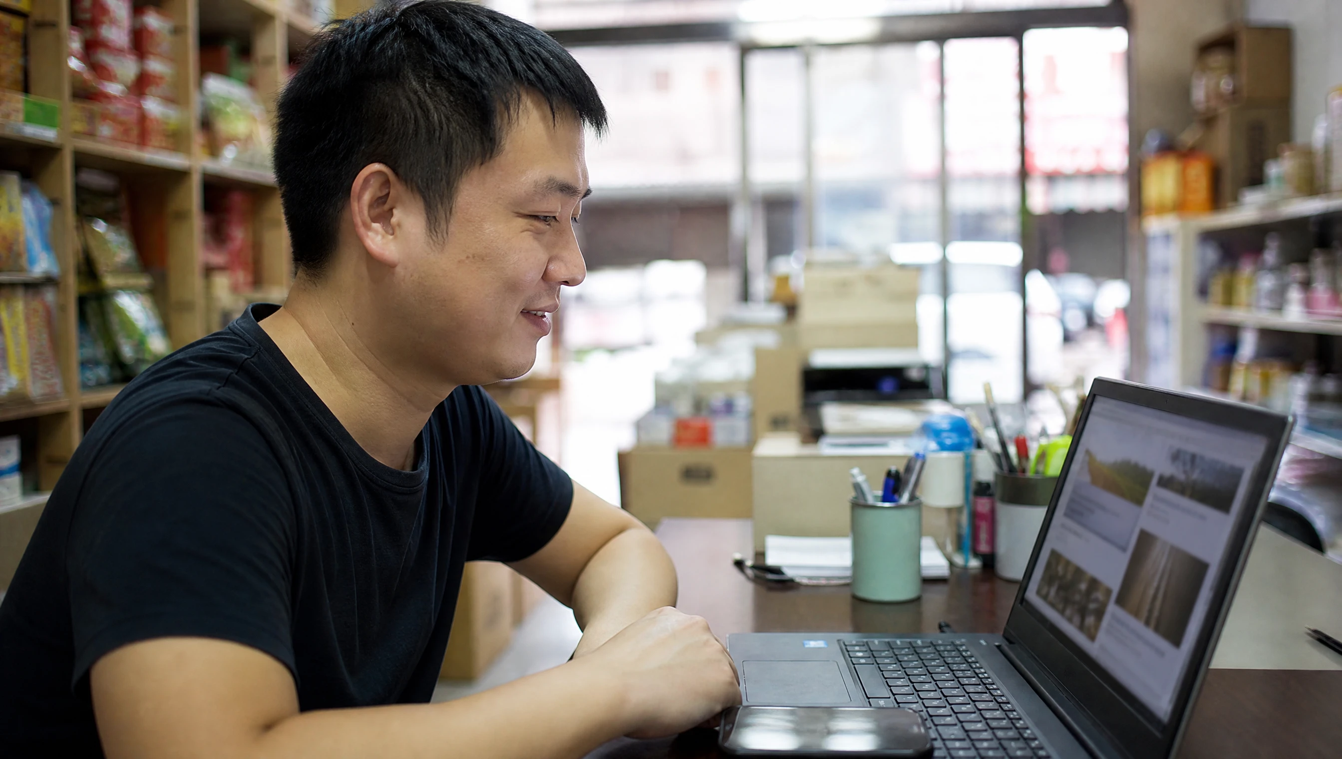 Malaysian business owner smiling while looking at his own website on a laptop in a shop
